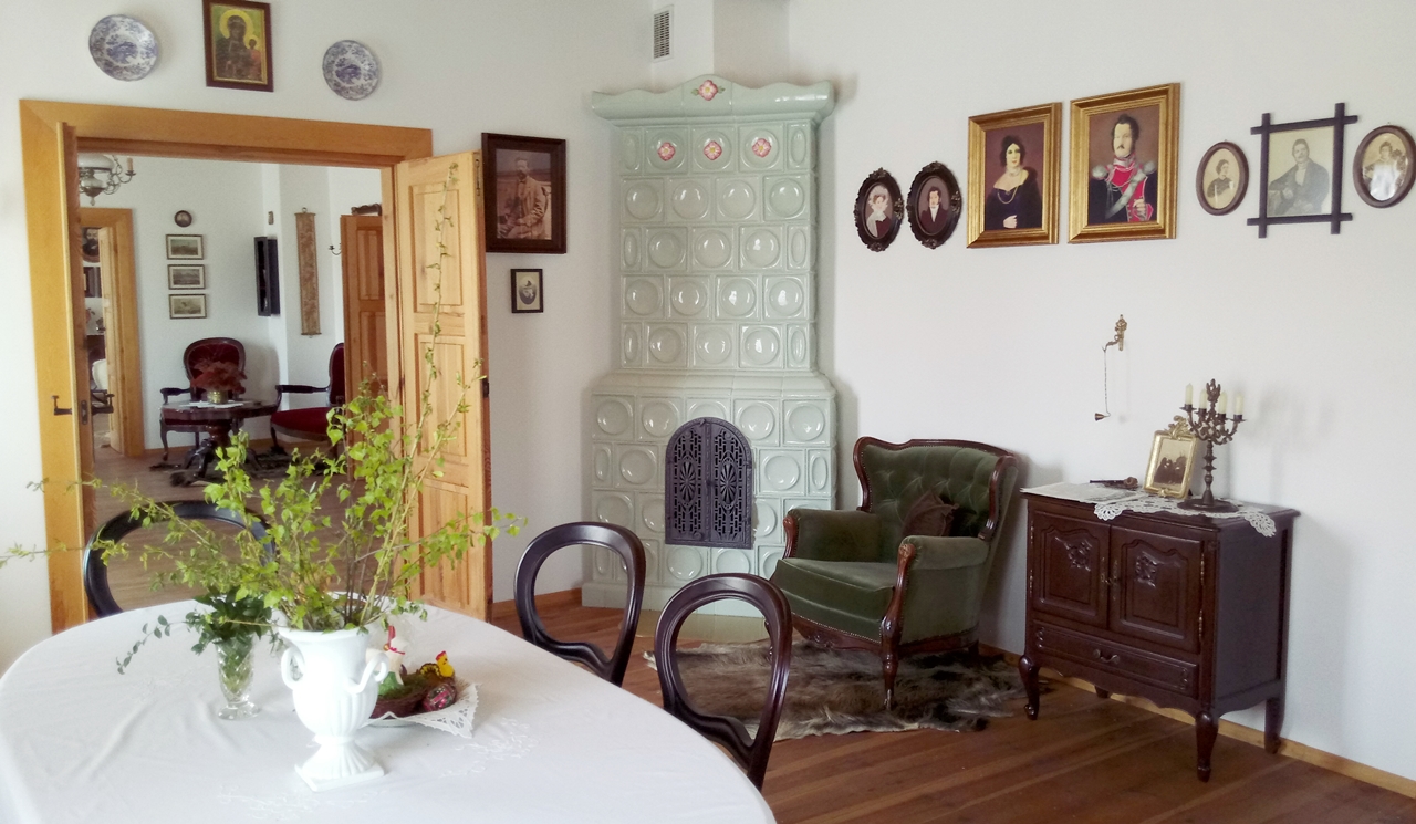 Salon z ceramicznym kominkiem w rogu, zielonym fotelem i stołem ukazujący dom rodzinny Stefana Żeromskiego, znajdujący się nieopodal Świętokrzyskiego Parku Narodowego.