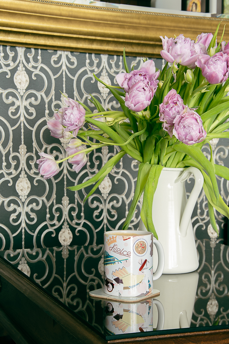 Kubek z charakterystycznymi grafikami dotyczącymi Kielc na tle wazonu z kwiatami.