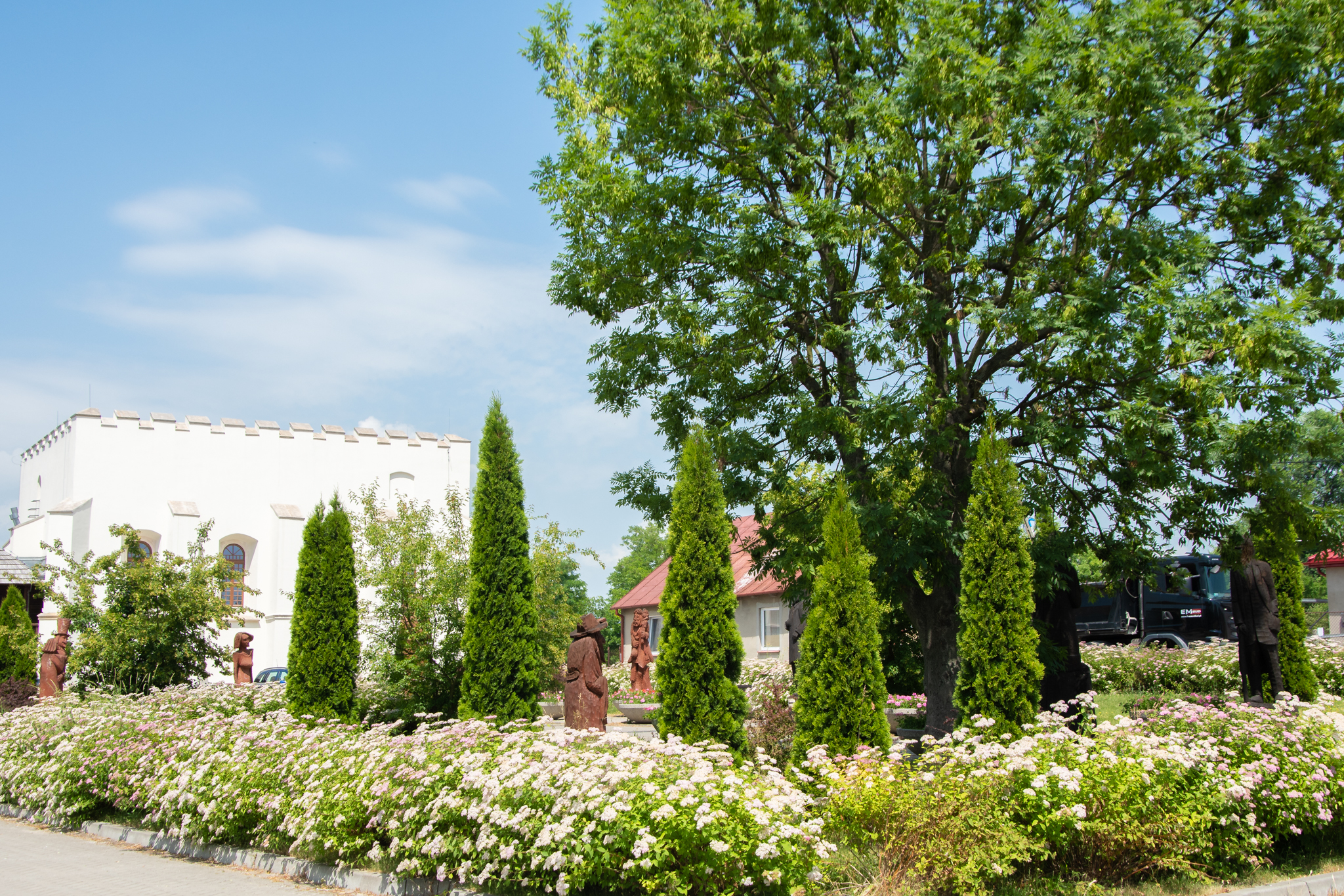 Biała, niewielka synagoga w kształcie kostki, jest schowana za kwiatami i małymi drzewkami.
