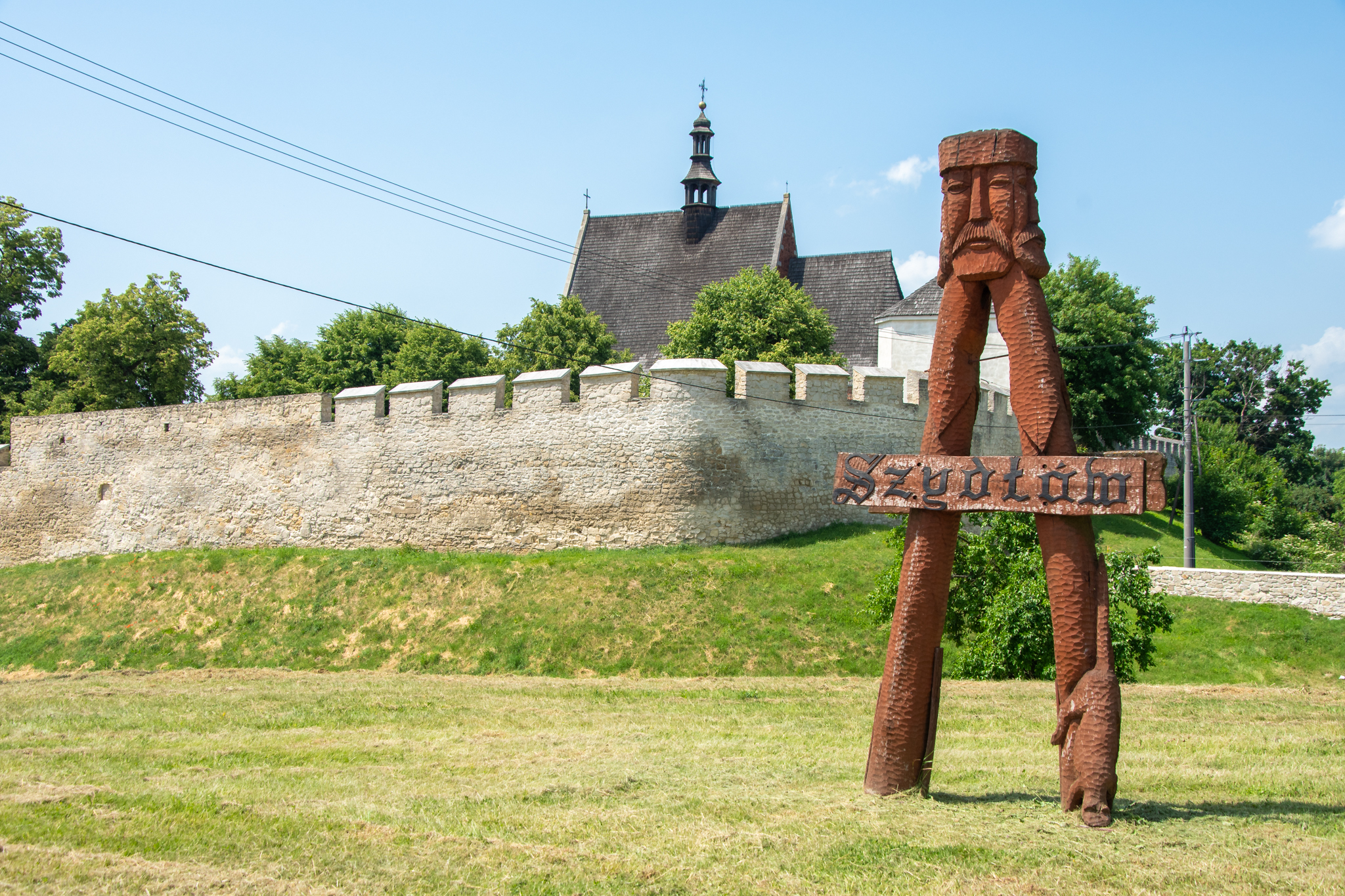 Drewniany znak z twarzą króla Kazimierza Wielkiego i znakiem "Szydłów", w oddali wskazuje na atrakcje Szydłowa - m.in. mury miejskie.