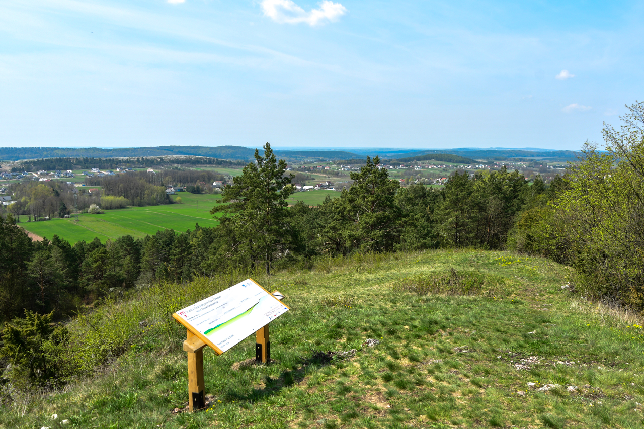 Szczyt Kamieniołomu Stokówka z widokiem na oddalone Góry Świętokrzyskie i tabliczkę turystyczną.