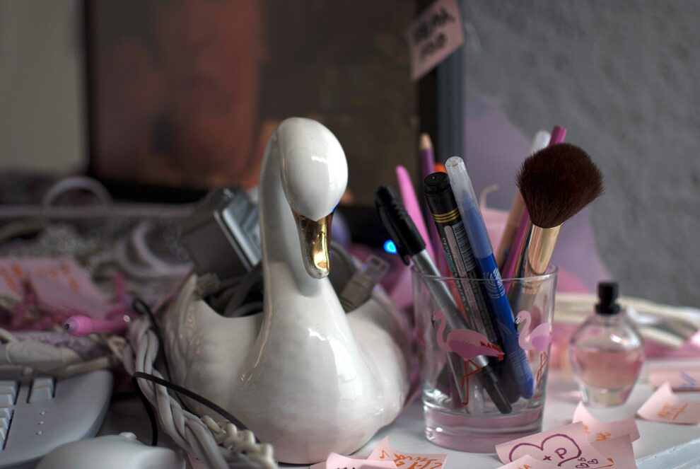 Szklana figurka łabędzia, obok szklanka z flamingami wypełniona markerami i pędzlami do makijażu. Motyw przypomina biurko influencera.